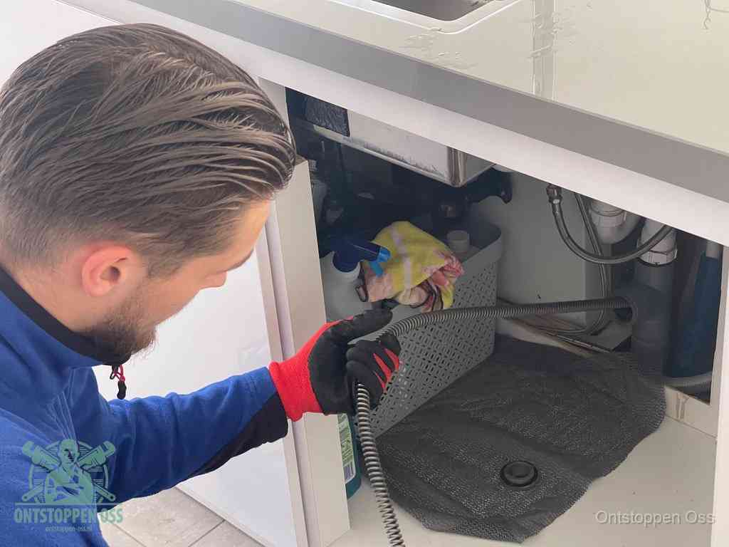 Een monteur voert een flexibele ontstoppingsveer in onder een keukenkastje om een verstopping voorbij de sifon te bereiken in Oss