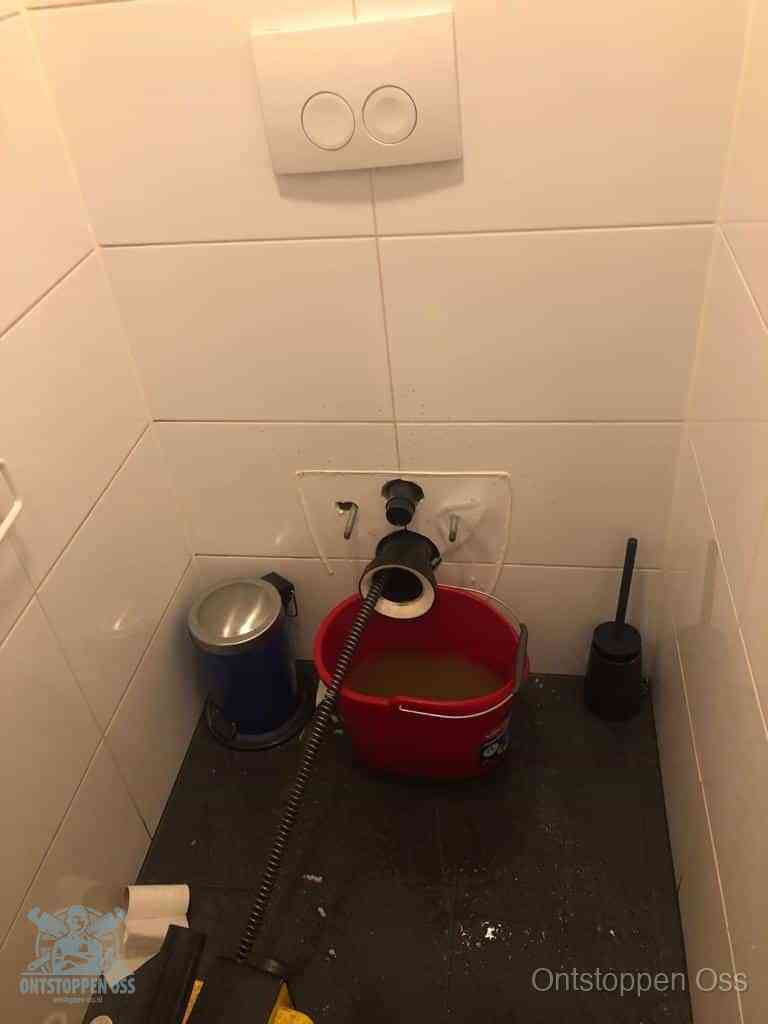 Een ontstopte wand-wc met verwijderde toiletpot waar een ontstoppingsveer in de wanduitgang verdwijnt, het achterhalen waar de verstopping zit in Oss