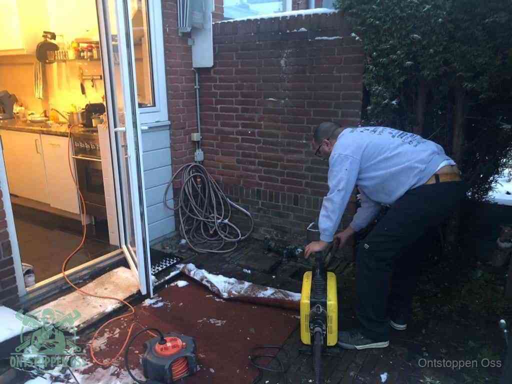 Een monteur werkt buiten bij een open riool-/grondput met een elektrische ontstoppingsmachine om een verborgen afvoerprobleem op te sporen in Oss