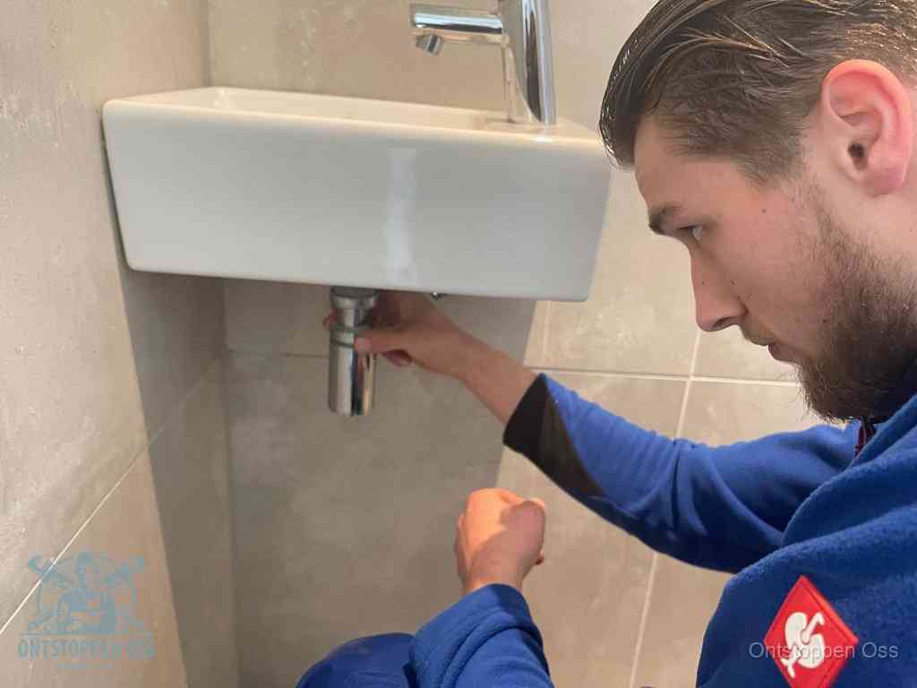 Een monteur draait met de hand de zwanenhalssifon onder een fonteinwasbak in de toiletruimte los om de bron van rioollucht in Oss te vinden