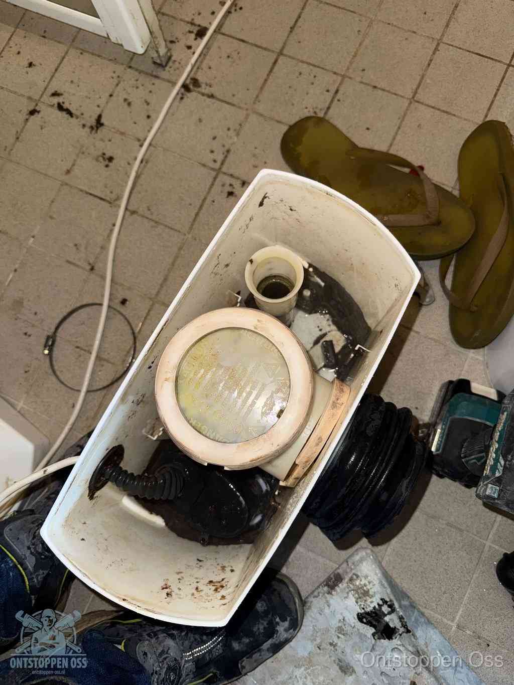 Een gedemonteerde toiletpot op de vloer met ontstoppingsveer en gereedschap ernaast, het verwijderen van een vastzittend voorwerp uit de wc in Oss
