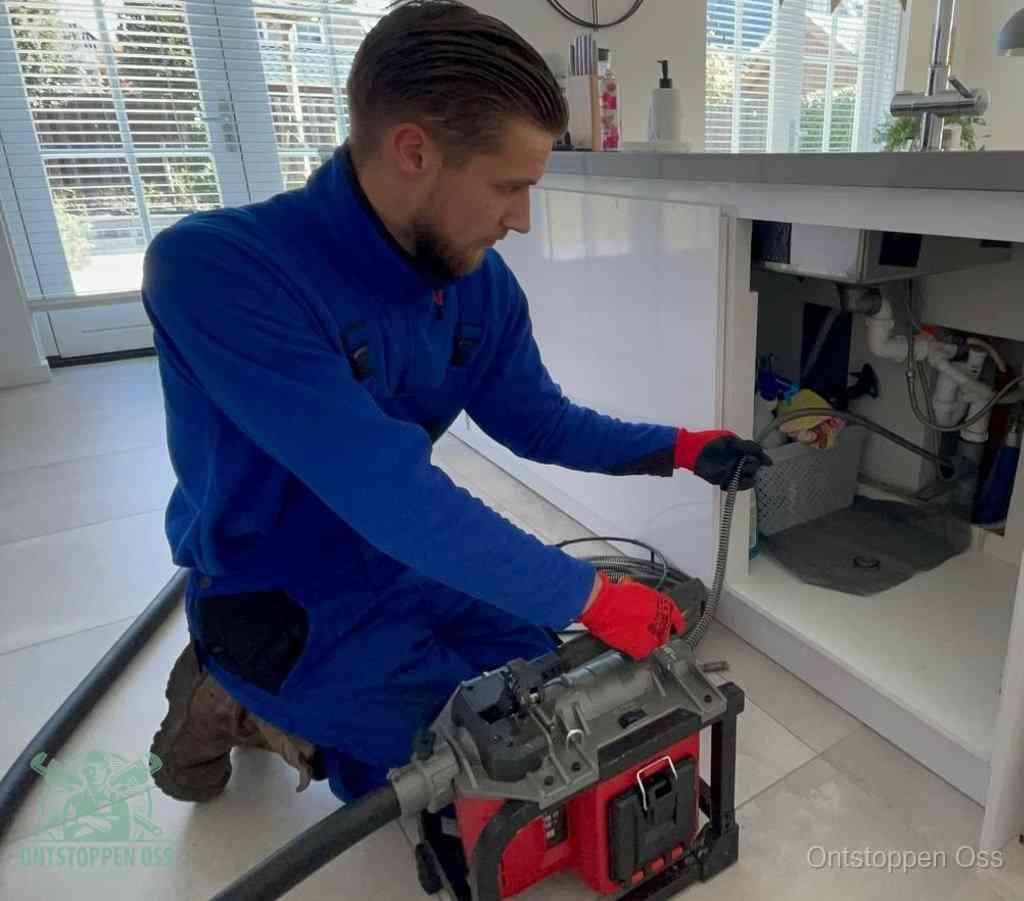 Een monteur met rode handschoenen knielt in een keuken en voert een elektrische ontstoppingsveer in tijdens het professioneel afvoer ontstoppen in Oss