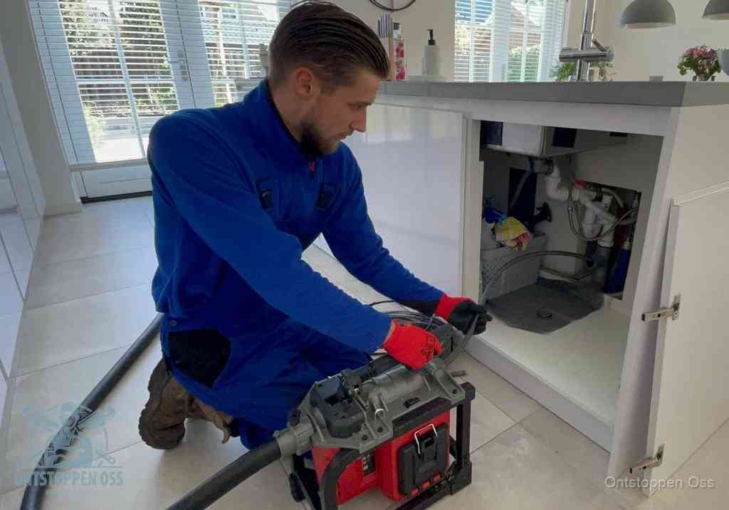 Een monteur voert een professionele elektrische ontstoppingsmachine in de afvoer onder de spoelbak tijdens het gootsteen ontstoppen in Oss